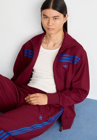 Tuta bordeaux realizzato in tessuto morbido, con strisce orizzontali blu e un piccolo logo. Il modello indossa una canotta bianca sotto.