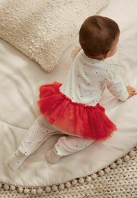 Hvid langærmet body med sorte prikker, parret med en rød tylskørt, båret over hvide leggings. Bløde teksturer mod en beige baggrund.