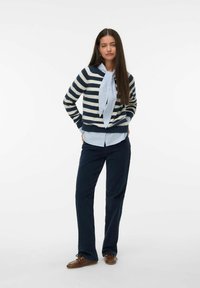 Pull rayé bleu marine et crème avec un détail de nœud papillon, superposé sur une chemise col bleu clair. Associé à un pantalon sombre et des chaussures marron.