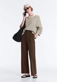 Femme portant une casquette beige, un t-shirt rayé à manches longues, un pantalon marron à jambes larges, des tongs noires, portant un sac noir, parlant sur un smartphone.