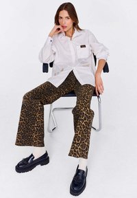 Biała koszula zapinana na guziki z kieszeniami, w połączeniu z brązowo-czarnymi spodniami w leopardzi wzór z dzwonami. Czarne buty z frędzlami i białe skarpetki.