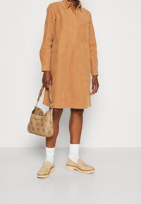 Braunes Cordhemdkleid mit langen Ärmeln und Knopfleiste. Beigefarbene Loafer und eine gemusterte beigefarbene Handtasche mit Schulterriemen. Weiße Socken.