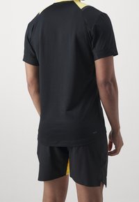 Camisa atlética negra con acentos amarillos, de manga corta y diseño texturizado. Combinada con pantalones cortos negros que tienen un panel lateral amarillo.