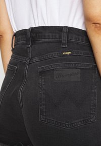 Shorts in denim nero a vita alta con orlo sfilacciato, dotati di una patch in pelle nera Wrangler, tasche posteriori e dettagli in oro nel logo.