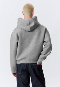 Pilkas gobtuvu megztinis, pagamintas iš minkštos, tekstūruotos medžiagos. Turi laisvą siluetą, nuleistus pečius ir ribines rankoves. Apačioje matyti tamsios džinsinės kelnės.