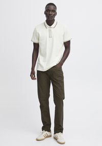 Polo en color blanco roto con cuello en contraste; combinado con pantalones verde oliva oscuro; modelo lleva zapatillas blancas con acentos marrones.
