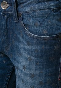 Mörkblå denimjeans med ett stjärnmönster i varierande nyanser. Inkluderar en metallknappstängning och traditionell fickdesign.