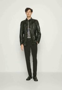 Černá kožená bunda s hladkým povrchem, dvěma předními kapsami, zapínáním na knoflík, nošená přes vzorovanou košili, spárovaná s úzkými černými kalhotami.