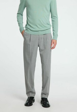 Graue, plissierte Hose mit seitlichen Taschen, aus glattem Stoff. Kombiniert mit einem hellgrünen Pullover und schwarzen Dressschuhen.