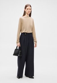 Beżowa bluzka z długim rękawem w połączeniu z ciemnymi, szerokimi spodniami z ozdobnymi złotymi guzikami. Modelka trzyma małą czarną torebkę. Gładka faktura tkaniny.
