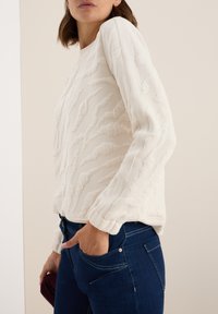 Pull en crème clair texturé avec des poignets et un ourlet côtelés, présentant des détails en relief, associé à un jean en denim foncé.