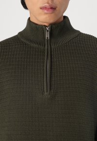 Pull à col zippé vert olive avec une texture côtelée. Il présente une demi-fermeture éclair et un col haut, mettant en valeur un design structuré.