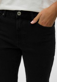 Mustat denim-šortsid, millel on vormitud disain, nähtav metallist nupp ja küljetaskud, sile ja veidi tekstureeritud kangas.