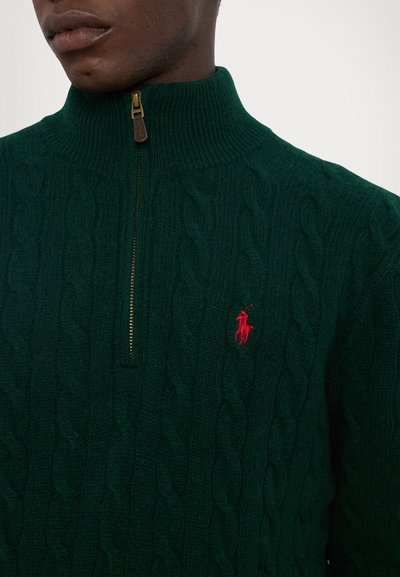 Dark green cable-knit sweater with a high collar and a half-zip closure, featuring a small red embroidered logo on the left chest.