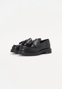 Mocasines de cuero negro con detalles de borlas, presentan una suela gruesa y texturizada con un diseño elegante. Superficie suave y forma de punta redonda.