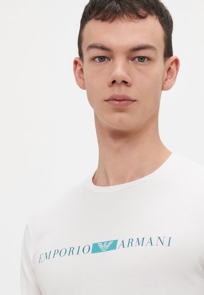 Balta trumpa rankovėmis marškinėliai su mėlynu Emporio Armani logotipu krūtinėje. Medžiaga atrodo lygi, su apvaliu kaklu.