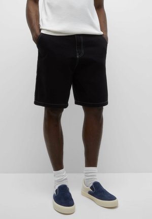 Persoon die zwarte shorts, witte sokken, blauwe instapschoenen en een wit T-shirt draagt, met handen in de zakken staand tegen een effen achtergrond.