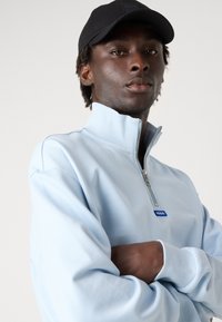 Sudadera de cremallera azul claro con cuello alto, puños acanalados y un parche de logo azul en el pecho. Una gorra negra completa el look.