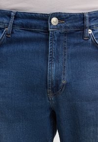 Jean en denim bleu foncé avec bouton et fermeture éclair à l'avant, passants de ceinture visibles et couture orange contrastante.