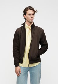 Jauns vīrietis ar viļņainiem matiem, ģērbies tumši brūnā jaka ar rāvējslēdzēju pār dzeltenu kreklu un gaiši zilām džinsām, skatās malā, ar roku kabatā.