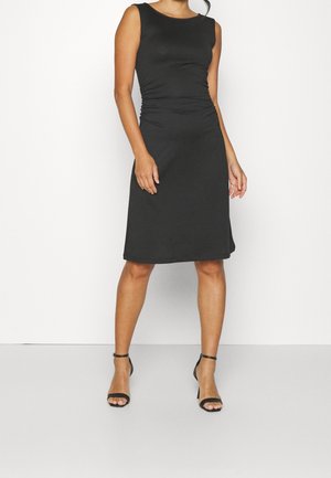 Kobieta w czarnej, bezrękawnikowej sukience do kolan z marszczeniem po bokach, nosząca czarne szpilki z paskiem wokół kostki, stoi na tle gładkiego, białego tła.