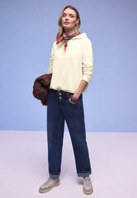 Vrouw staat met hand in de zak, draagt een lichtgele hoodie, rode sjaal met patroon, opgerolde blauwe jeans, witte sneakers, en houdt een bruine jas vast.