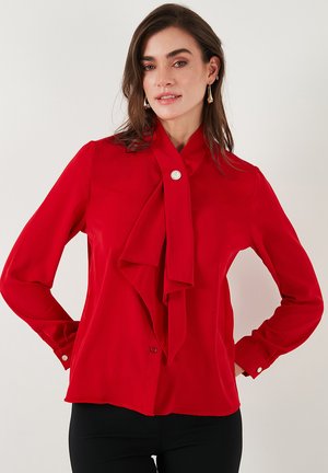 Red blouse made of smooth fabric featuring a tied bow at the collar, long sleeves with buttoned cuffs, and a single button closure at the neck.