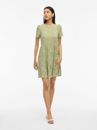 Vestito verde a maniche corte con design a strati, caratterizzato da un motivo a pois bianchi. Realizzato in tessuto leggero e abbinato a sandali open-toe.