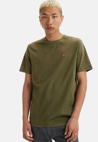 Ung person med kort, blond hår iført en olivengrøn Levi's t-shirt og mørke jeans, stående foran en hvid baggrund.