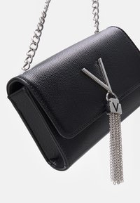Pochette en cuir texturé noir avec une chaîne argentée, présentant un insigne géométrique et une breloque en métal. Design rectangulaire compact.