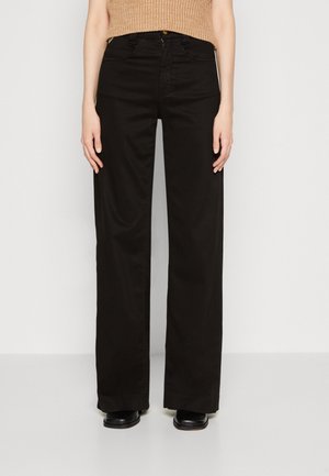 Osoba ubrana w czarne, szerokie spodnie z wysokim stanem, beżowy bezrękawnik dzianinowy oraz czarne buty, stojąca na tle jednolitego, jasnego tła.