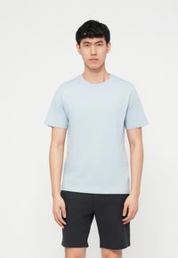 Νεαρός άνδρας που στέκεται, φοράει ένα ανοιχτό γαλάζιο κοντομάνικο μπλουζάκι και μαύρα σορτς, μπροστά από ένα απλό λευκό φόντο.