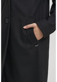 Cappotto nero realizzato in tessuto texturizzato, con chiusura a bottoni, tasca laterale e un accento argentato del marchio vicino all'orlo.