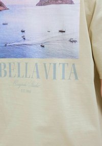 Šviesus smėlio spalvos marškinėliai su fotografiniu laivų ant vandens atspaudu, papildyti užrašu „BELLA VITA“ ir švelnaus, lygios tekstūros.
