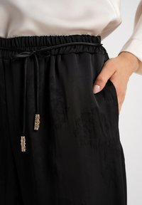 Czarne spodnie z elastycznym pasem, wyposażone w troczek z metalicznymi akcentami oraz subtelną kwiatową fakturą. Widoczna boczna kieszeń.