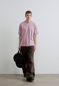 Svetloružové bavlnené tričko s okrúhlym výstrihom a detailom loga, v kombinácii s voľnými hnedými nohavicami a čiernou športovou taškou.