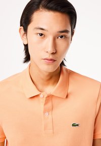 Polo de color naranja claro con una tela texturizada, que cuenta con una botonera de tres botones y un logo de cocodrilo verde en el lado izquierdo del pecho.