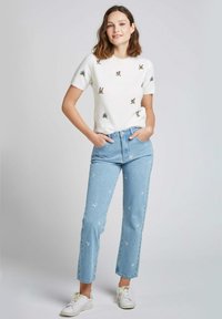 Pull court à manches courtes blanc avec broderies florales, assorti à un jean en denim bleu clair avec des motifs floraux blancs, et des baskets blanches.