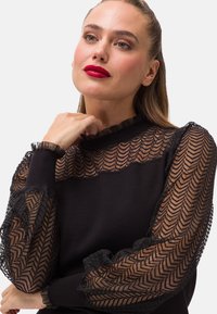 Mustapullo, jossa on teksturoitu pitsihihat, joissa on herringbone-kuvio, kerätyt hihansuut ja ribattua kaula-aukko. Sujuva kangasrunko.