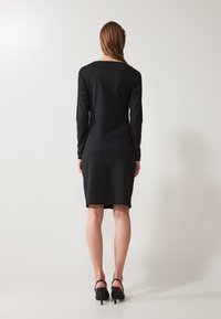 Robe noire à manches longues avec un design ajusté, longueur genou, texture lisse et décolleté arrondi. Portée avec des chaussures à talons hauts noires.