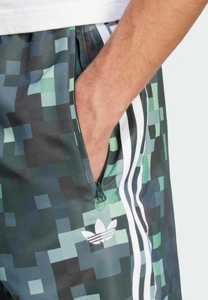 Hand in pocket of green and blue pixelated patterned shorts with white stripes and Adidas logo.