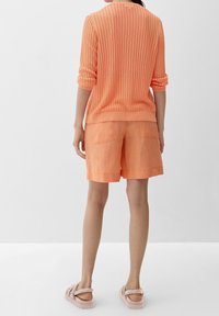 Pomarańczowy sweter z prążkowanym wzorem w zestawie z dopasowanymi pomarańczowymi lnianymi szortami. Miękka tekstura, podwinięte rękawy i tylnie kieszenie w szortach. Jasne sandały.