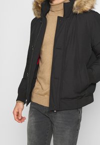 Schwarze Pufferjacke mit gefütterter Kapuze aus Kunstfell, Reißverschluss- und Knopfverschluss sowie elastischen Bündchen. Getragen über einem beigen Pullover und grauen Jeans.
