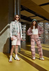 Två modeller poserar inomhus. Den ena har på sig en pastellfärgad pufferväst över en vit skjorta, mönstrade rosa shorts och unika sneakers. Den andra har en rosa bomberjacka, vit topp och batikmönstrade rosa jeans.