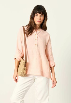 Frau trägt eine hellrosa Bluse mit Knöpfen und hochgekrempelten Ärmeln, weiße Hose, eine beige gewebte Schultertasche und eine bunte Perlenkette.