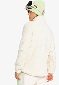 White faux fur jacket with a high collar and long sleeves, paired with a light green fuzzy beanie and ski goggles on the forehead.