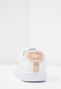 White sneaker with a smooth leather upper and a beige textured heel patch featuring an embroidered logo. White rubber sole, minimalistic design.