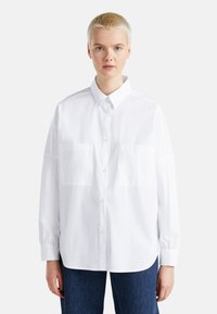 Camicia bianca con colletto, due tasche frontali, maniche lunghe e vestibilità comoda. Realizzata in tessuto leggero con una texture liscia.