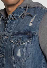 Denim vest med en grå hættetrøje, med slidte detaljer, metal knapper og to brystlommer. Blå denim kontrasterer med det grå stof.