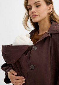 Cappotto leggero marrone con un ampio cappuccio e due bottoni. Presenta una tasca frontale che contiene un oggetto bianco e peloso con una parte superiore tonda.